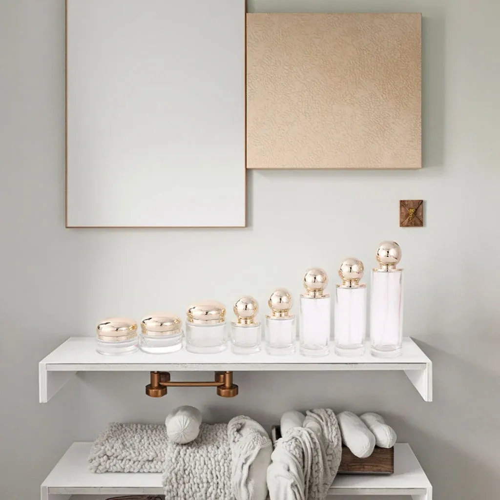 Clear glass skincare set with gold ball lids styled on a bathroom shelf with towels