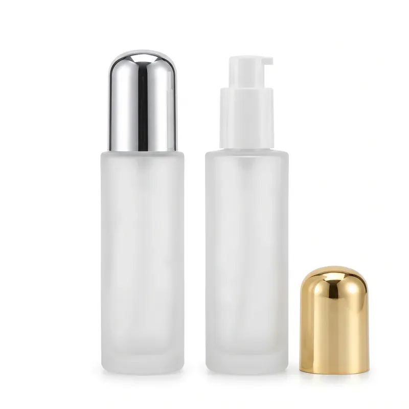 Two frosted glass cylindrical pump bottles on white background: one with a polished silver dome overcap, one with the white lotion pump exposed and its gold dome overcap resting beside