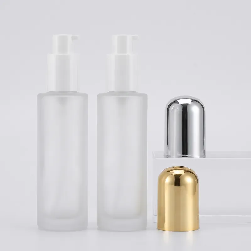 Frosted glass lotion pump bottles with white pumps exposed, with a polished silver dome overcap on a clear acrylic riser and the gold dome overcap standing on the surface in front