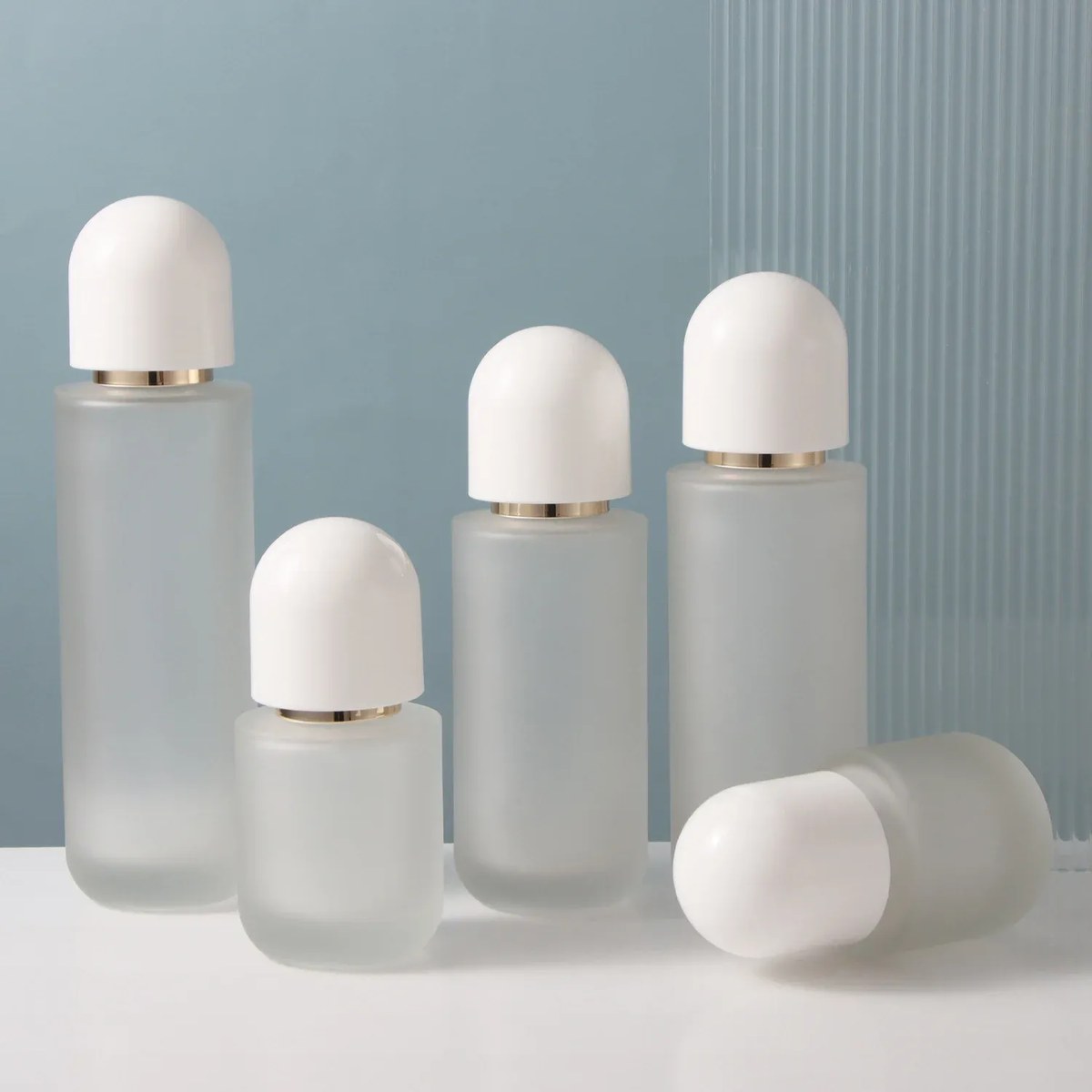 Frosted glass skincare set with white dome caps: four bottles and a lying bottle on a blue-grey background