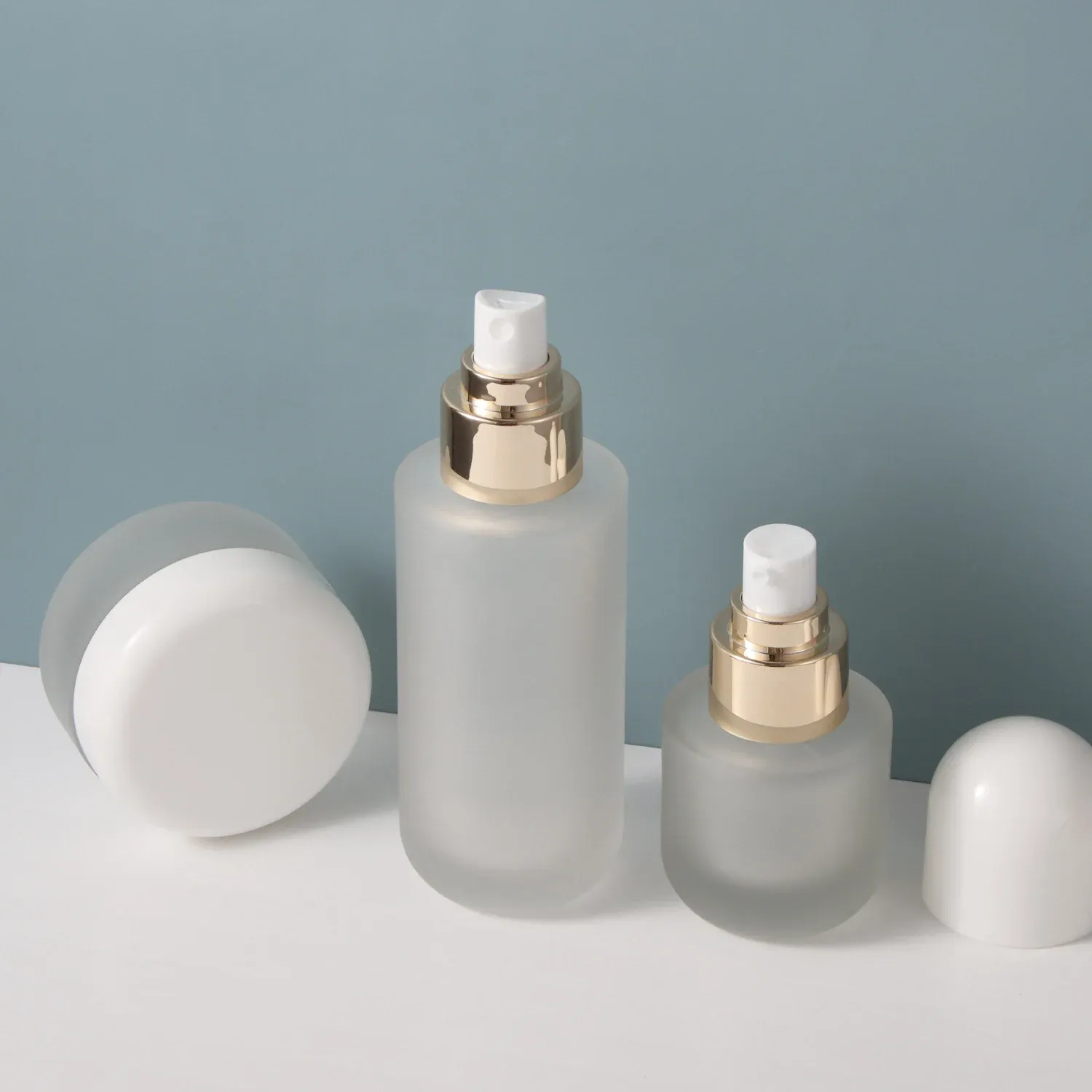 Frosted glass bottles with gold pumps and white actuator, dome cap removed, cream jar beside