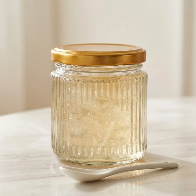 A single squat ribbed glass jar filled with translucent brewed bird's nest with its gold lid on, ceramic spoon beside