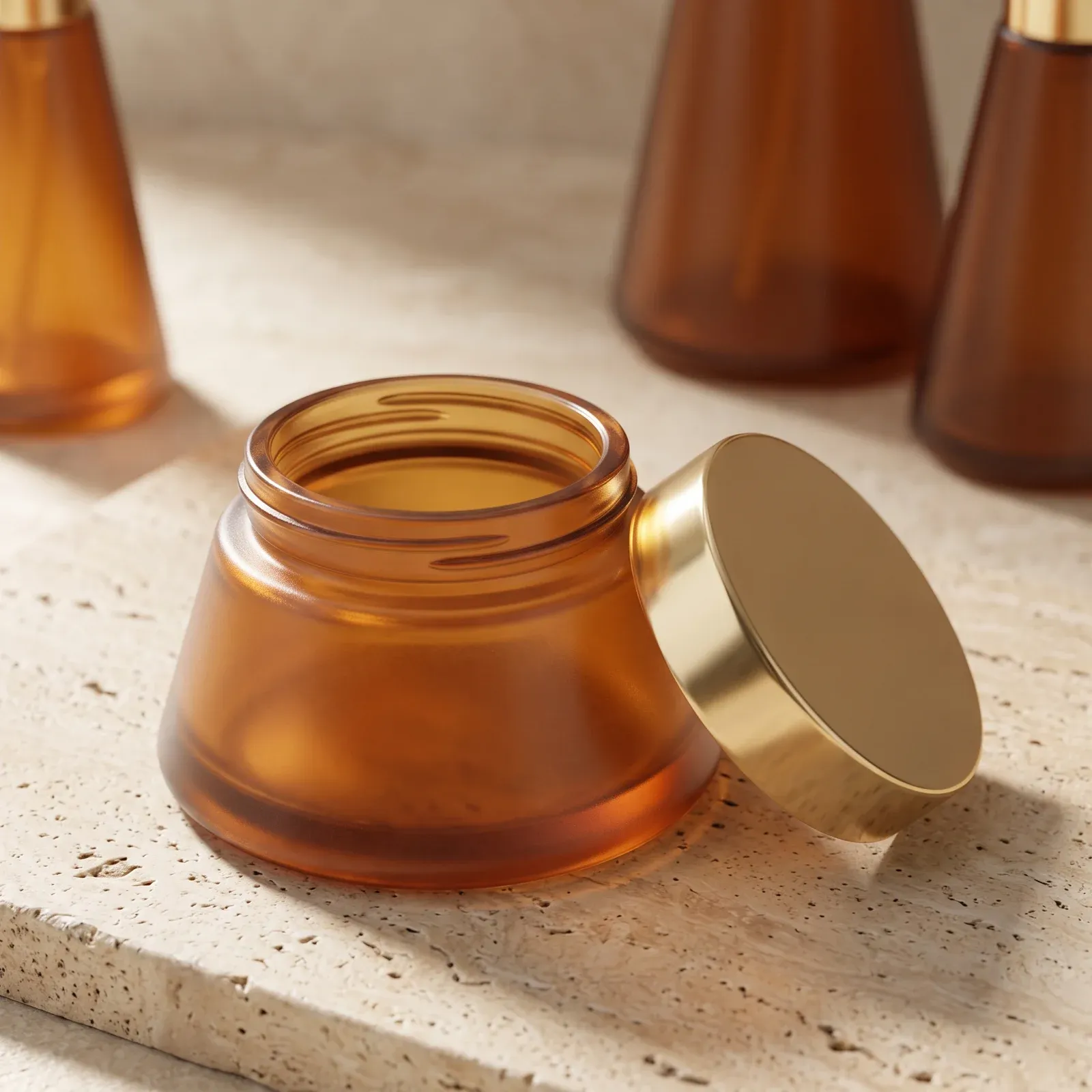 Open amber conical glass cream jar with its flat gold lid leaning beside it on travertine stone