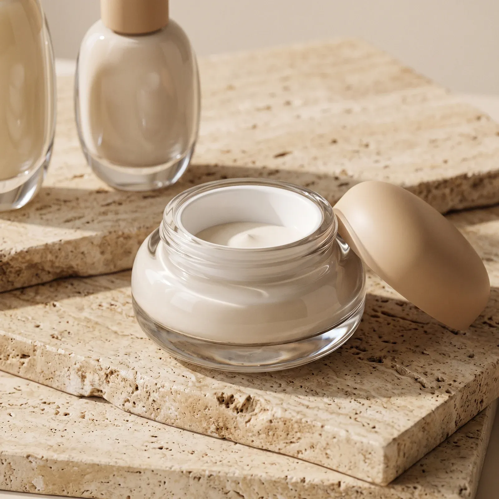 Open pebble-shaped glass cream jar with white inner liner and its sand-tone dome lid beside it